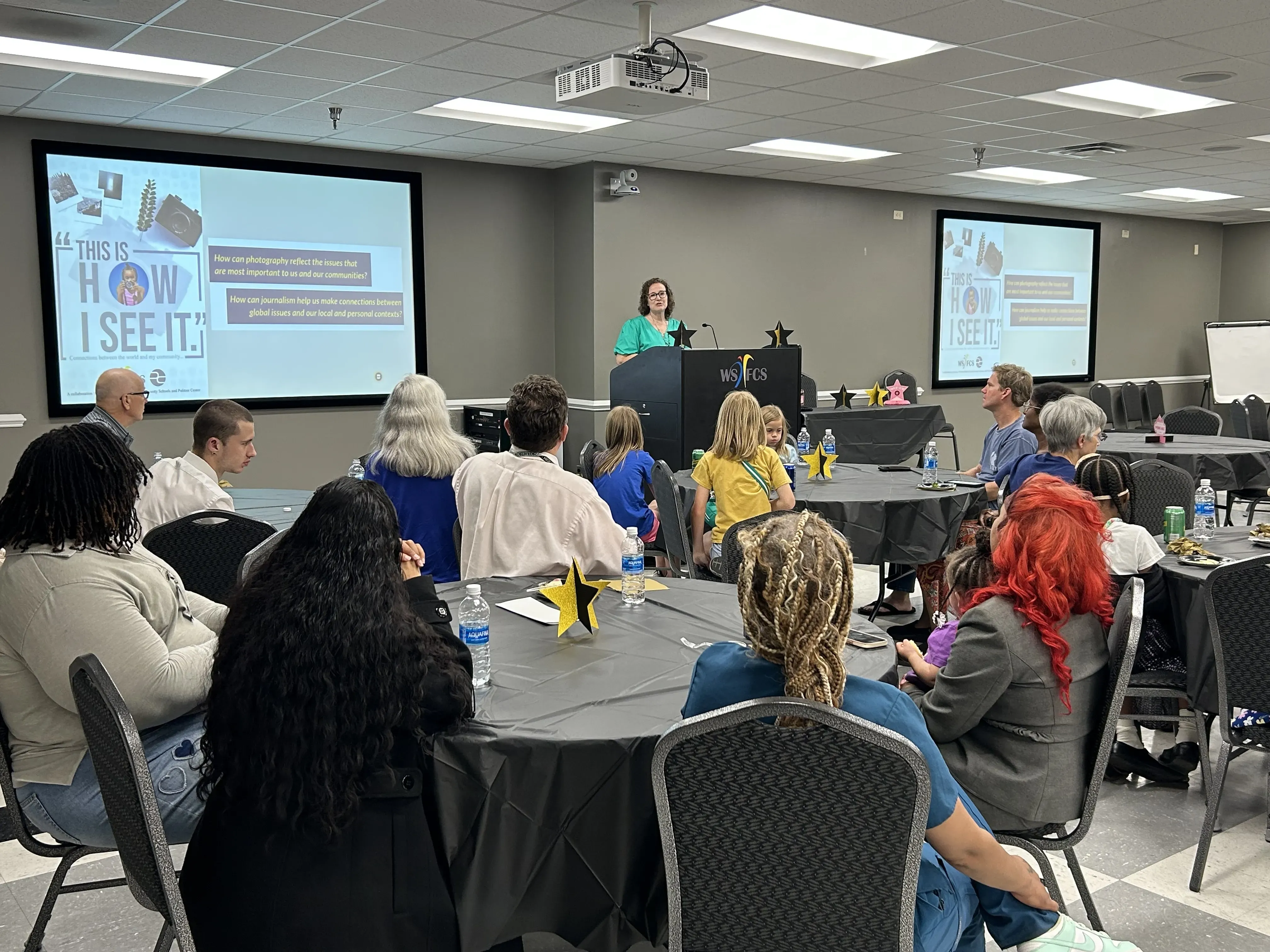 Students and their families sit at tables looking at a screen with the cover image for the How I See It photo contest. A woman stands at a podium in front of the screen and speaks to the students.
